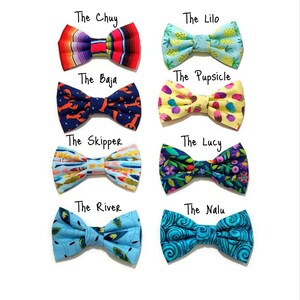 May include: Eight different bow ties with various colorful patterns, including pineapples, lobsters, fish, and swirls. Each bow tie has a name written above it: The Chuy, The Lilo, The Baja, The Pupsicle, The Skipper, The Lucy, The River, and The Nalu.
