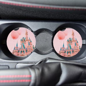 May include: Two pink car coasters with a pink castle and pink balloons. The coasters are in a car cup holder.