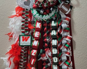 May include: A decorative homecoming mum in red, white, and green, featuring ribbons, feathers, and a teddy bear. The mum includes the names "KENZIE" and "LANDON" in white letters, along with a football and the year "2021".