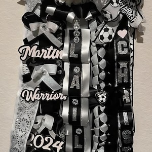 Peut inclure: Une décoration de bal de promo avec un ballon de football. Elle présente des rubans noirs, blancs et argentés, avec les noms "Martin" et "Lila". Le mot "Warriors" et l'année "2024" sont également visibles.