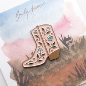 May include: A pink enamel pin featuring a floral cowboy boot with blue flowers and green leaves. The pin is on a card with a watercolor background and the text "Emily June".