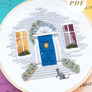 May include: An embroidery hoop containing a stitched house facade with a bright blue door, a gold heart-shaped knocker, two windows with yellow and purple curtains, grey brickwork, floral bushes, and a small grey cat sitting on the front step.