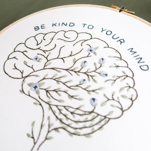 Pode incluir: Arte de bordado em aro com um design de c&eacute;rebro estilizado, rodeado de folhas verdes e flores azuis. O texto "Be kind to your mind" &eacute; bordado em fio azul.
