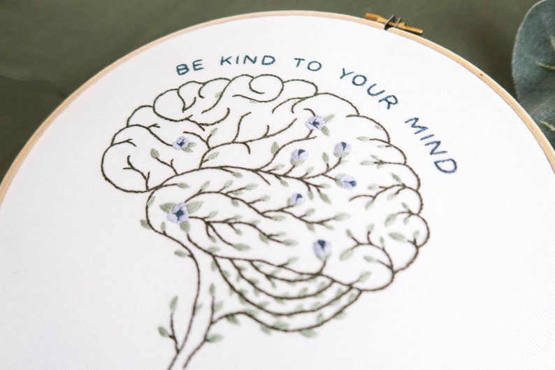 Pode incluir: Arte de bordado em aro com um design de c&eacute;rebro estilizado, rodeado de folhas verdes e flores azuis. O texto "Be kind to your mind" &eacute; bordado em fio azul.