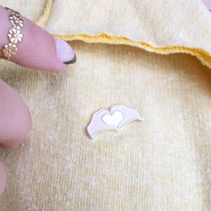 Pu&ograve; includere: Uno spillo in smalto dorato con due mani che formano un cuore con un cuore bianco al suo interno. Lo spillo &egrave; su uno sfondo di tessuto giallo.
