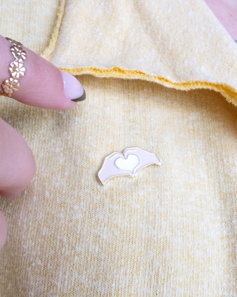 Pu&ograve; includere: Uno spillo in smalto dorato con due mani che formano un cuore con un cuore bianco al suo interno. Lo spillo &egrave; su uno sfondo di tessuto giallo.