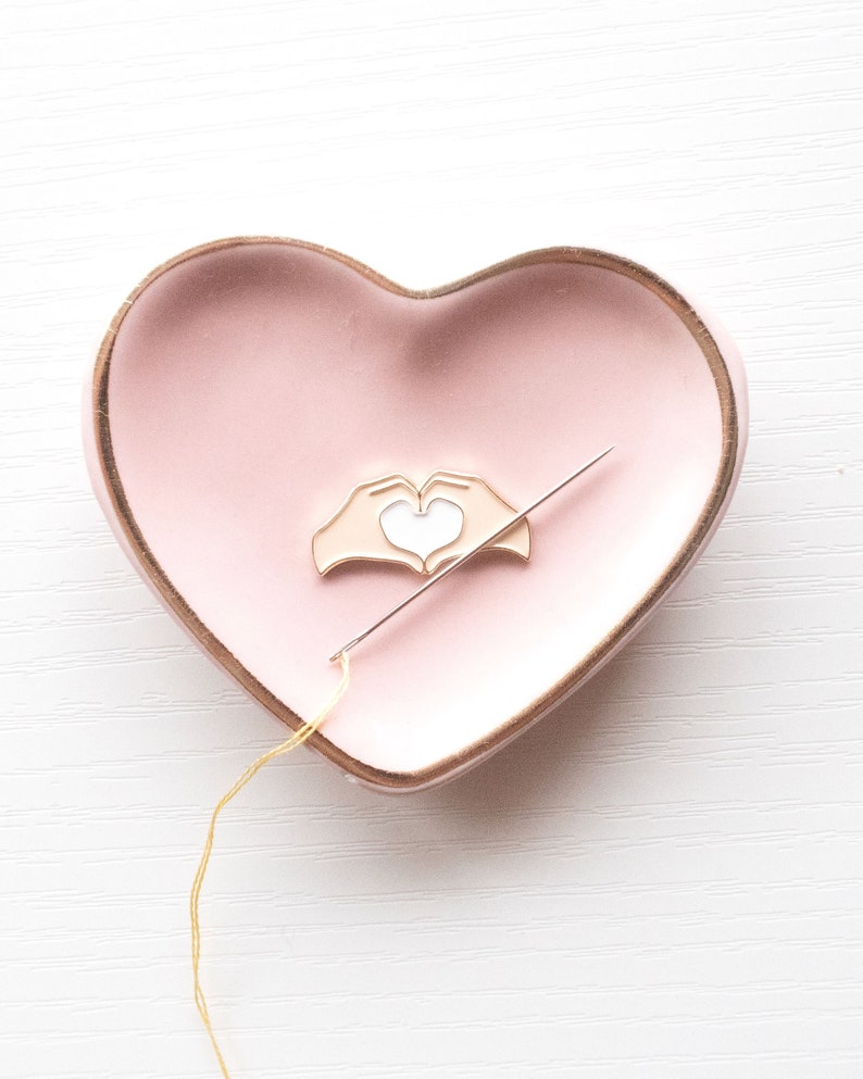 Pu&ograve; includere: Uno spillo a forma di cuore dorato con un cuore bianco all'interno, appoggiato su un piatto a forma di cuore rosa. Un filo dorato &egrave; infilato in un ago, che &egrave; anche appoggiato sul piatto.