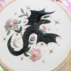 May include: A circular wooden embroidery hoop containing white fabric with a hand-stitched design of a dark, shimmering dragon surrounded by light pink flowers, green leaves, a silver crescent moon, and small metallic gold star accents.