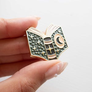 May include: A gold-colored enamel pin in the shape of an open book. The pin features a dark green cover with white stars and a crescent moon. The pages are light pink, and the spine is black and brown.