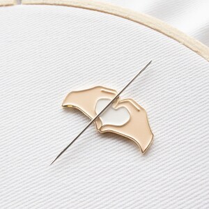 Pu&ograve; includere: Uno spillo in metallo color oro con un design di due mani che formano una forma di cuore. Le mani sono color pesca e il cuore &egrave; bianco. Lo spillo &egrave; su uno sfondo di tessuto bianco.
