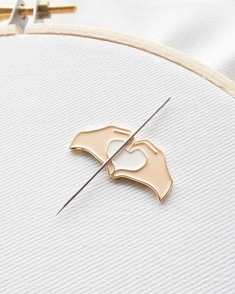 Pu&ograve; includere: Uno spillo in metallo color oro con un design di due mani che formano una forma di cuore. Le mani sono color pesca e il cuore &egrave; bianco. Lo spillo &egrave; su uno sfondo di tessuto bianco.