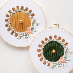 May include: Two embroidery hoops with white fabric and wooden frames. The embroidery designs are of a round door with a gold or green center and brown flower accents. Each door has a gold bead in the center.