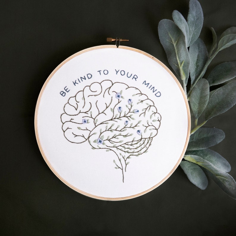 Pode incluir: Aro de bordado branco com um c&eacute;rebro bordado em azul e verde com folhas e flores. O texto "Be kind to your mind" &eacute; bordado em torno do c&eacute;rebro.