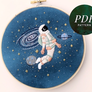 Op de afbeelding: Borduurwerk in een hoepel met een astronaut die in de ruimte zweeft met een sterrenstelsel, sterren en planeten. De astronaut draagt een wit ruimtepak met een groen en roze ontwerp. De achtergrond is een donkerblauwe stof.