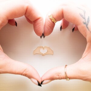 Pu&ograve; includere: Due mani che formano un cuore con un ciondolo a forma di cuore dorato a forma di mano al centro. Le mani hanno lo smalto nero e un anello d'oro sull'indice della mano destra.