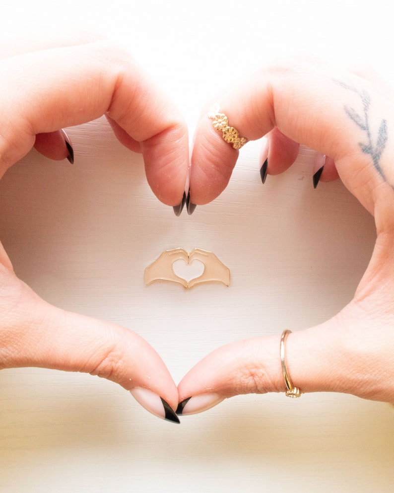 Pu&ograve; includere: Due mani che formano un cuore con un ciondolo a forma di cuore dorato a forma di mano al centro. Le mani hanno lo smalto nero e un anello d'oro sull'indice della mano destra.