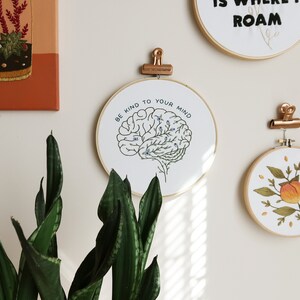 Pode incluir: Aro de bordado branco com moldura dourada, apresentando um contorno preto e branco de um c&eacute;rebro com folhas verdes e flores azuis. O texto "Be kind to your mind" est&aacute; escrito em um c&iacute;rculo ao redor do c&eacute;rebro.