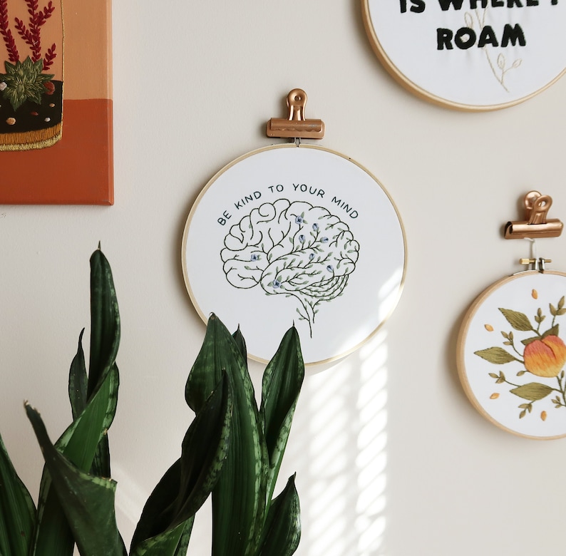 Pode incluir: Aro de bordado branco com moldura dourada, apresentando um contorno preto e branco de um c&eacute;rebro com folhas verdes e flores azuis. O texto "Be kind to your mind" est&aacute; escrito em um c&iacute;rculo ao redor do c&eacute;rebro.