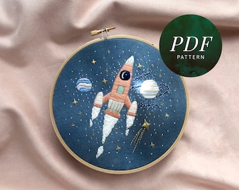 May include: Embroidered hoop art featuring a rocket ship flying through a starry night sky with planets. The rocket is orange and white with a blue flame. The planets are blue and white with gold rings. The background is a dark blue fabric with white stars.