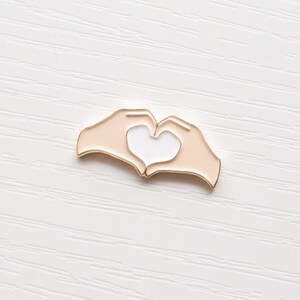Pu&ograve; includere: Spilla in smalto dorata con due mani che formano una forma di cuore. Le mani sono color pesca con un cuore bianco al centro.