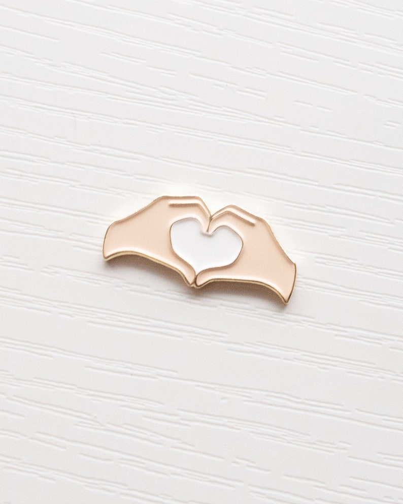 Pu&ograve; includere: Spilla in smalto dorata con due mani che formano una forma di cuore. Le mani sono color pesca con un cuore bianco al centro.