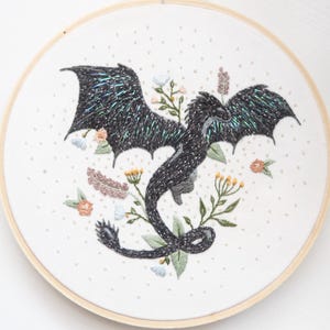 Puede incluir: Bordado de arte de un dragón negro con alas iridiscentes, rodeado de flores bordadas en tonos melocotón, amarillo y azul, en un aro de madera. El fondo blanco está salpicado de pequeños puntos blancos.
