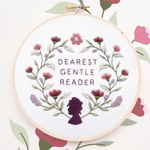 May include: A circular wooden embroidery hoop containing white fabric with the stitched text DEAREST GENTLE READER in dark purple. The text is framed by a floral wreath of pink and purple flowers with green leaves and a dark purple silhouette.