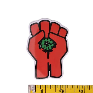 Puede incluir: Pegatina de puño rojo con un ojo verde en la palma. La pegatina mide aproximadamente 5 cm de alto.
