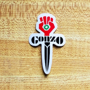 May include: A white sticker featuring a black dagger with the word "GONZO" in black lettering. The dagger has a red fist with a green four-leaf clover at the top.