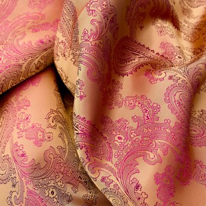 K&ouml;nnte beinhalten: Nahaufnahme eines Stoffes mit Paisley-Muster. Der Stoff ist hellbeige mit einem pink- und lilafarbenen Paisley-Muster.