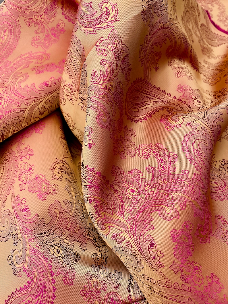 K&ouml;nnte beinhalten: Nahaufnahme eines Stoffes mit Paisley-Muster. Der Stoff ist hellbeige mit einem pink- und lilafarbenen Paisley-Muster.