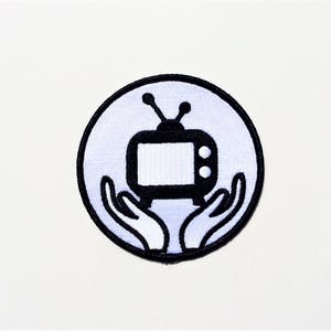 May include: A white circle with a black outline, featuring a black and white television with antennas and two hands holding it up. The television has a white screen and two black knobs.
