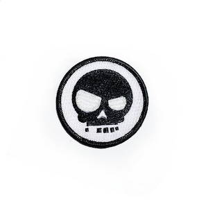 May include: Black and white embroidered patch with a skull design. The skull is black with white eyes and a white mouth. The patch is surrounded by a black circle.