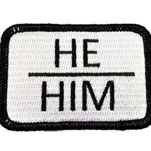 May include: White fabric patch with black embroidered text that reads "HE/HIM".