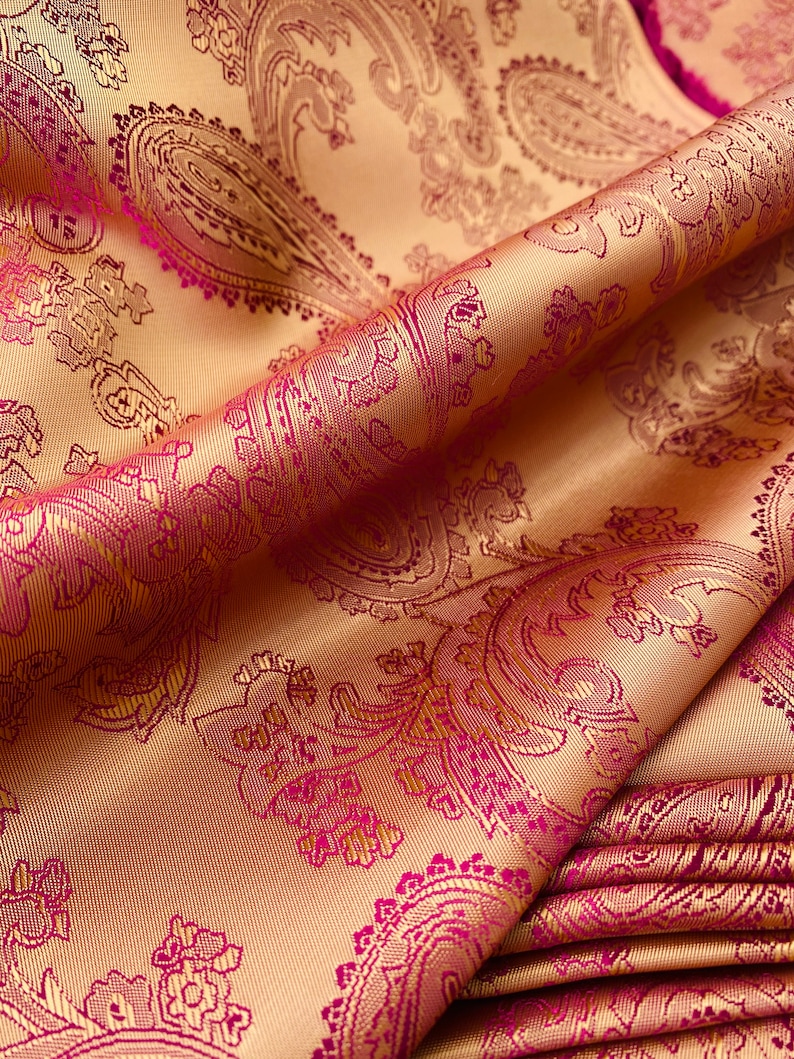 K&ouml;nnte beinhalten: Nahaufnahme eines Stoffes mit Paisley-Muster. Der Stoff ist hellgoldfarben mit einem tiefrosa Paisley-Muster.