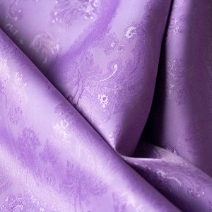 May include: A close-up of a soft, purple fabric with a white paisley pattern. The fabric is folded and draped, showing its texture and drape.