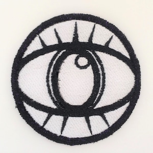 以下が含まれることがあります： 黒い刺繍で目のデザインが施された白い布のパッチ。目は太い黒い輪郭と中央に小さな黒い円があります。目は黒いまつげの輪で囲まれています。