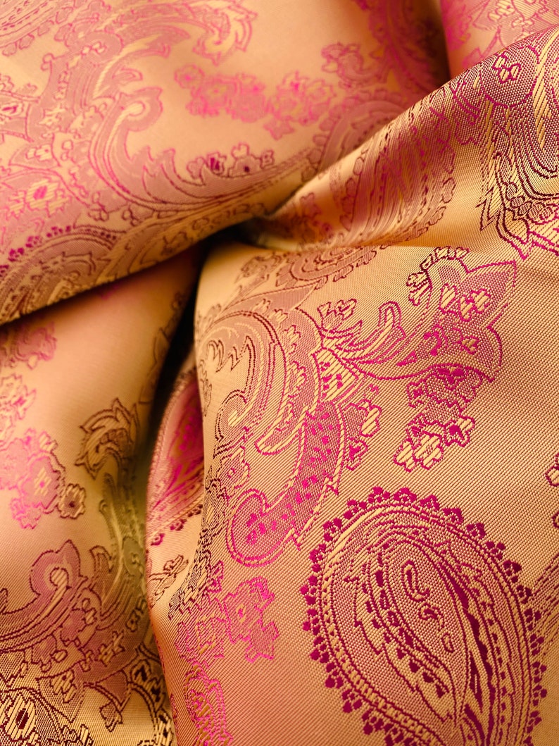 K&ouml;nnte beinhalten: Nahaufnahme eines Stoffes mit Paisley-Muster. Der Stoff ist hellgoldfarben mit einem rosafarbenen Paisley-Muster.