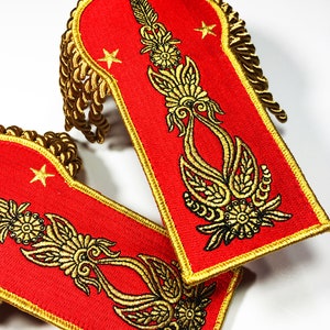May include: Two red fabric shoulder patches with gold embroidered trim and a gold embroidered design. The design features a stylised floral motif with a star at the top.