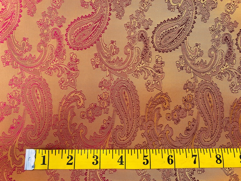 K&ouml;nnte beinhalten: Nahaufnahme eines Stoffes mit Paisley-Muster in Rosa- und Goldt&ouml;nen. Ein gelbes Ma&szlig;band liegt auf dem Stoff und zeigt die Ma&szlig;e von 1 bis 9 Zoll.