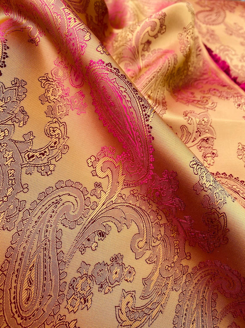 K&ouml;nnte beinhalten: Nahaufnahme eines goldenen Stoffes mit einem rosafarbenen Paisley-Muster. Der Stoff ist weich und luxuri&ouml;s, und das Muster ist filigran und detailliert.