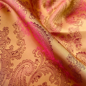 Op de afbeelding: Een close-up van een stof met een paisley-patroon in roze en gouden tinten. De stof is zacht en luxueus, met een subtiele glans.