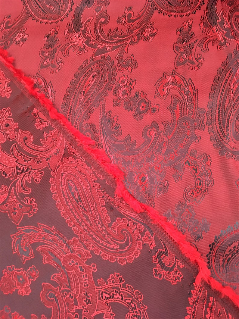 May include: Red paisley fabric with a black and red pattern. The fabric has a textured surface and a fringed edge.