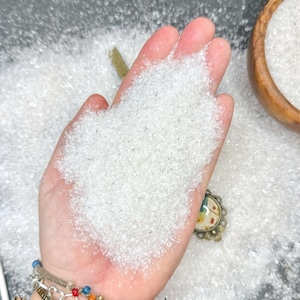 Könnte beinhalten: Eine Handfläche gefüllt mit einem Haufen feiner, durchscheinender weißer Kristallkörnchen. Die Textur wirkt körnig und glitzernd, ähnlich wie grobes Salz oder Zucker. Ein kleiner Teil eines dekorativen Anhängers mit Blumenmuster ist sichtbar.