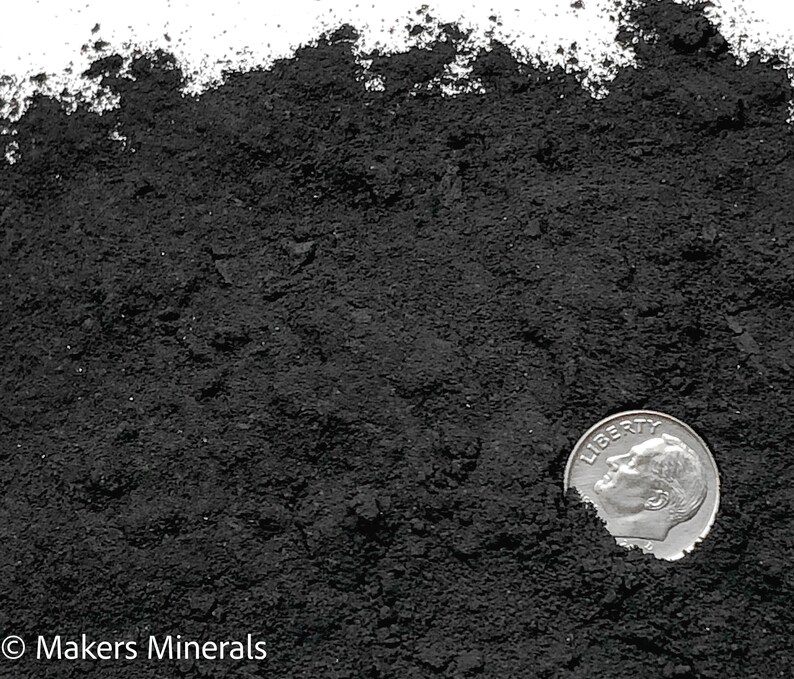 May include: A pile of fine, matte black mineral powder with a coarse, granular texture. A silver-coloured United States dime coin is partially embedded in the lower right side of the powder for scale. The text Makers Minerals appears in the bottom left.