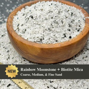 May include: A wooden bowl filled with Rainbow Moonstone and Biotite Mica sand. The sand is a mix of white, black, and iridescent particles. The text "Rainbow Moonstone + Biotite Mica" and "Coarse, Medium, & Fine Sand" is displayed.