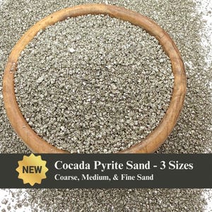 May include: A wooden bowl filled with pyrite sand, with a dark gray background. The sand is a mix of coarse, medium, and fine grains. The text on the image reads "Cocada Pyrite Sand - 3 Sizes" and "Coarse, Medium, & Fine Sand."