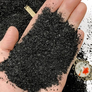 Op de afbeelding: Een hoopje kleine, grove, zwarte minerale korrels in een handpalm. De textuur is onregelmatig en korrelig, vergelijkbaar met steengruis of houtskool. Een deel van een goudkleurige metalen liniaal en een bloemvormige hanger zijn zichtbaar.