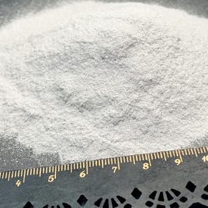 May include: A pile of fine, light gray powder with a granular texture. A black ruler is positioned at the bottom of the frame, showing measurements in inches, with the visible scale ranging from 4 inches to 10 inches across the bottom edge.