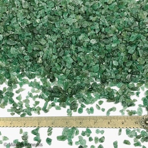 May include: A collection of small, irregular, translucent green mineral fragments scattered across a surface. A metal ruler is positioned horizontally at the bottom, showing measurements in centimeters from 1 to 10. The text Makers Minerals is visible.